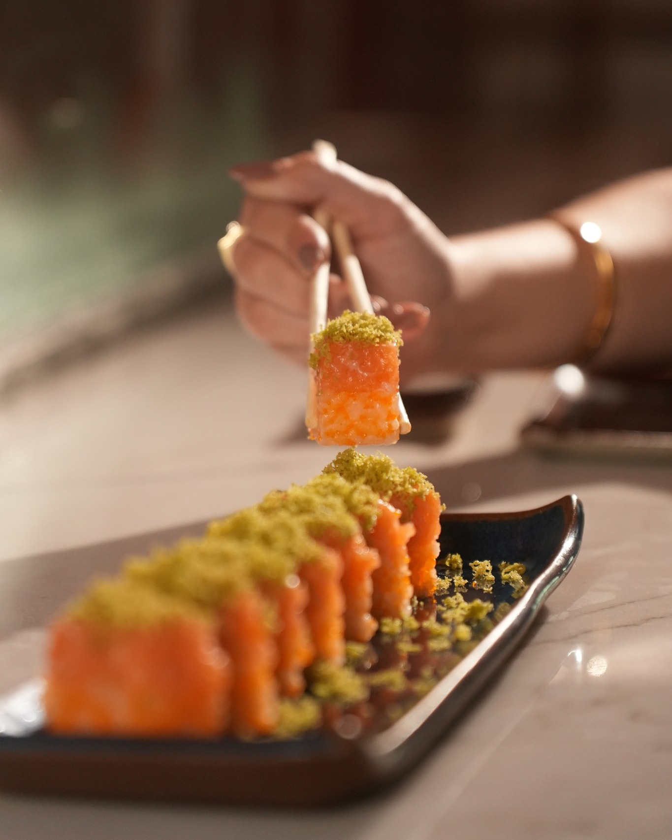 Convidada segurando com hashi uma peça de uramaki de salmão finalizada com farofa crocante verde, prato completo em primeiro plano