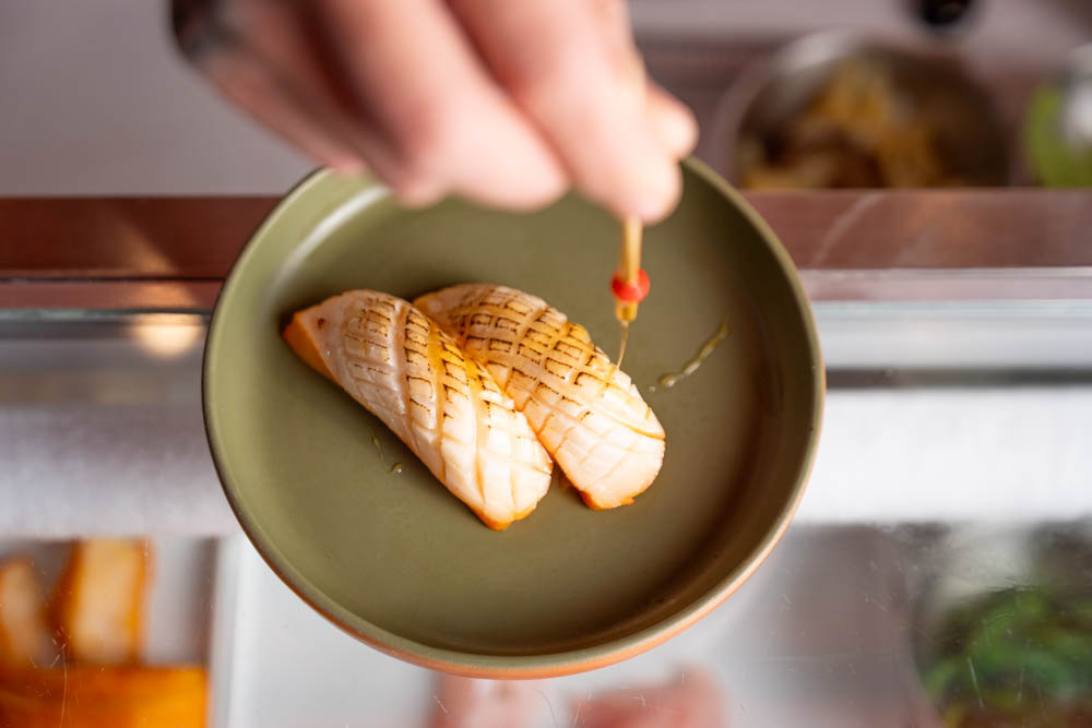 Duas peças de nigiri maçaricado recebendo fio de mel em prato verde sage