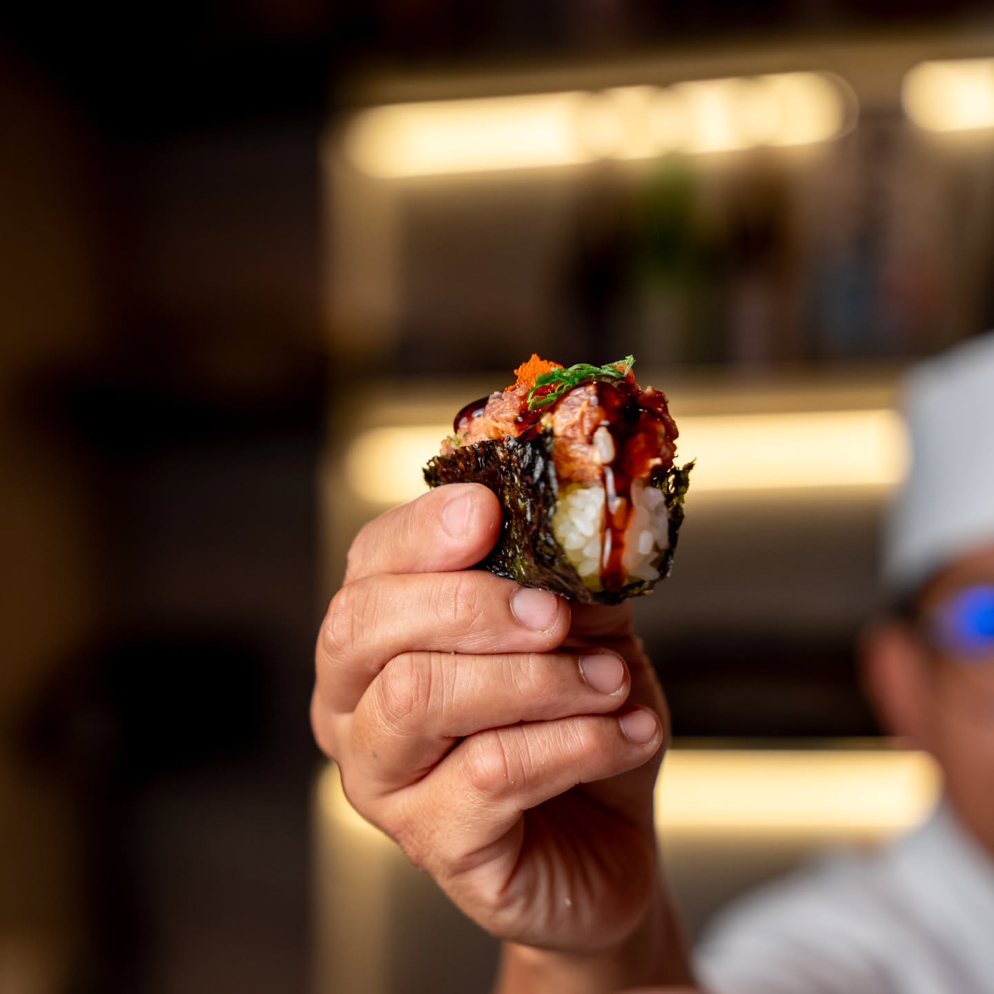 Mão do chef apresentando um gunkan de tartare de salmão com ovas alaranjadas, cebolinha e molho tarê