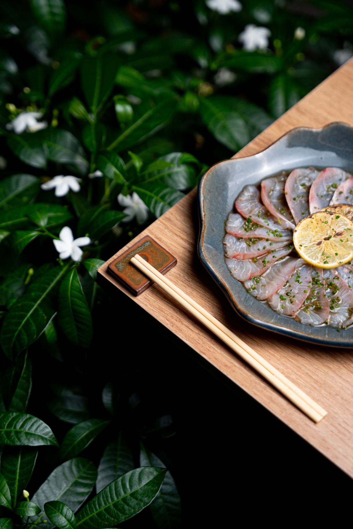Prato de peixe cru com limão tostado, hashi e fundo de folhas verdes com flores brancas